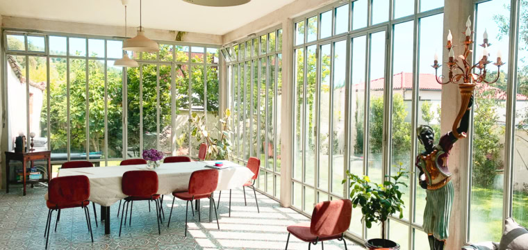 Salle ensoleillée avec table blanche, chaises rouges et plantes décoratives.