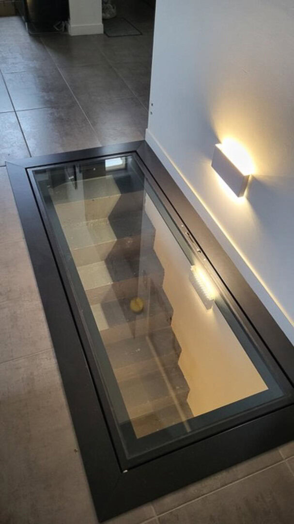 Trappe de sol vitrée en métal, révélant un escalier menant à un niveau inférieur, conçue et installée par un métallier rhônalpin.