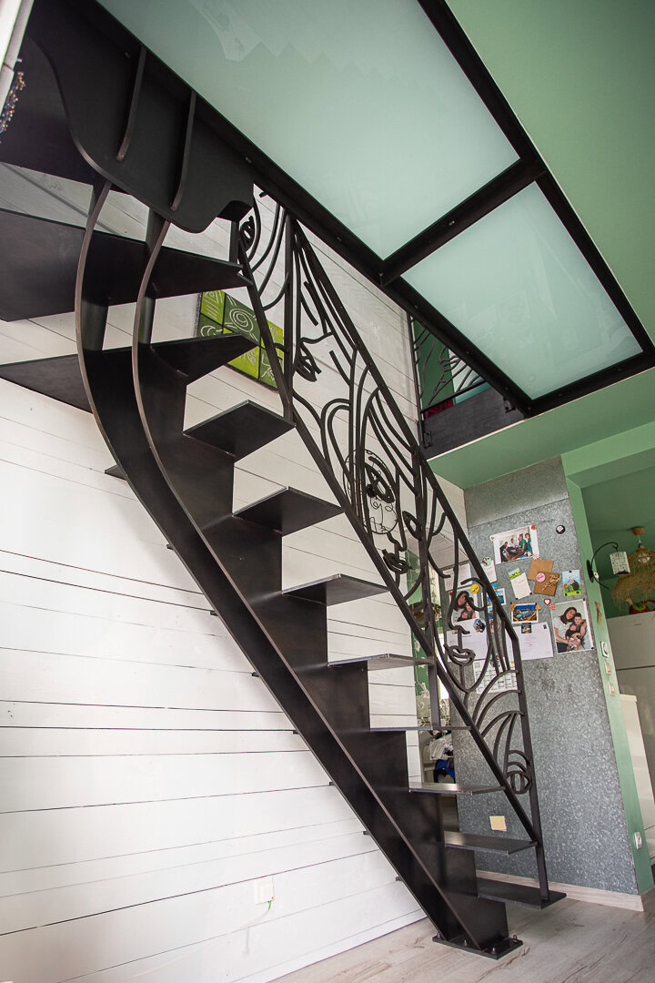 Escalier en métal noir avec rampe décorative, mur en panneaux blancs et plancher en verre au-dessus, réalisé par un métallier lyonnais.